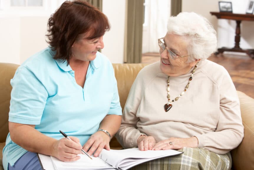 Senior Woman In Discussion With Health Visitor At Home Writing Down Information Smiling At Each Other